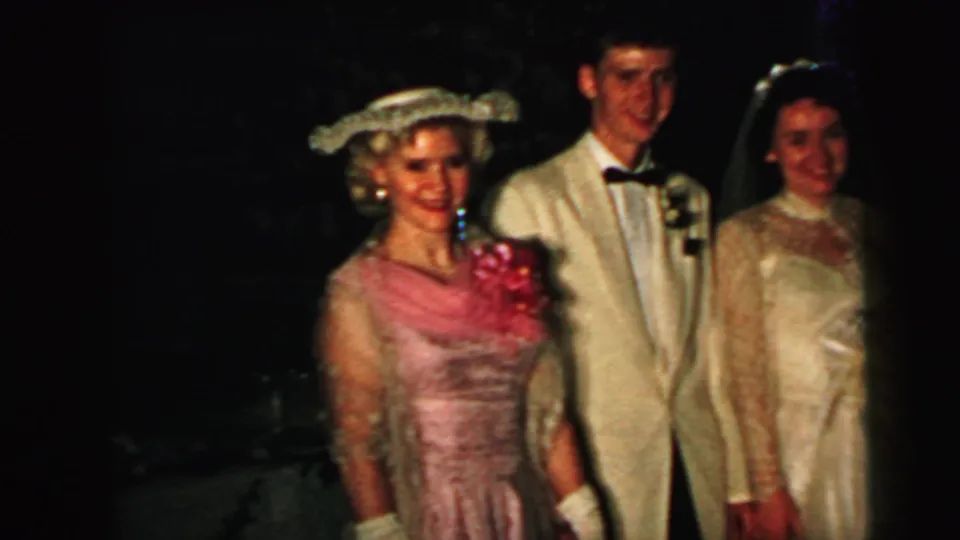 1964 Camden wedding frame with a pink-dressed guest, tuxedoed man, and bride smiling in a dark interior.
