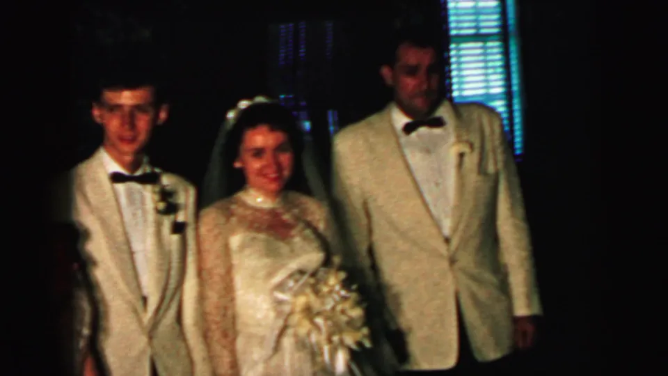 1964 Camden home-movie frame showing a bride between two men in cream formal jackets at a wedding.