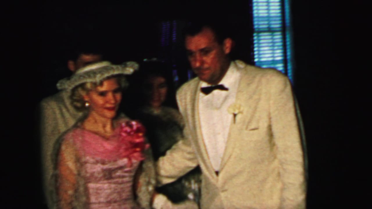 Bride in pink-toned dress and hat walks beside a man in a cream tuxedo in a dim interior.