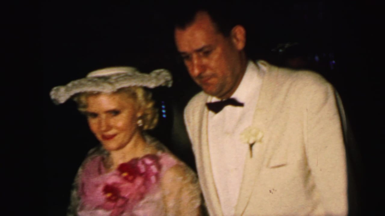1964 Camden wedding footage shows a woman in pink formalwear and a white hat near a man in a cream tuxedo under dim indoor light.