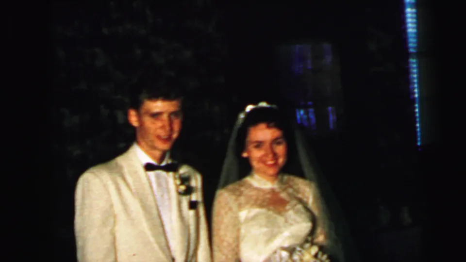 In 1964 Camden wedding footage, a bride in a lace gown and veil stands beside a groom in a white jacket and black bow tie.