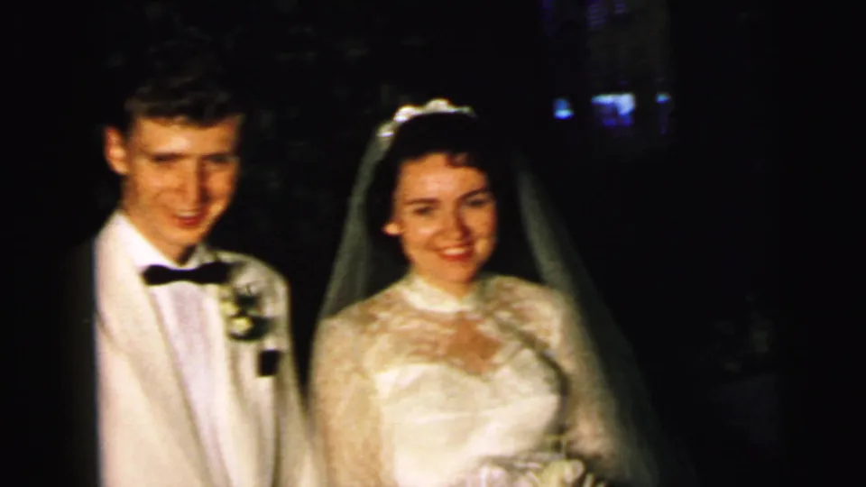 A closer 1964 home-movie frame shows the groom in a white jacket beside the bride in a lace gown and veil.