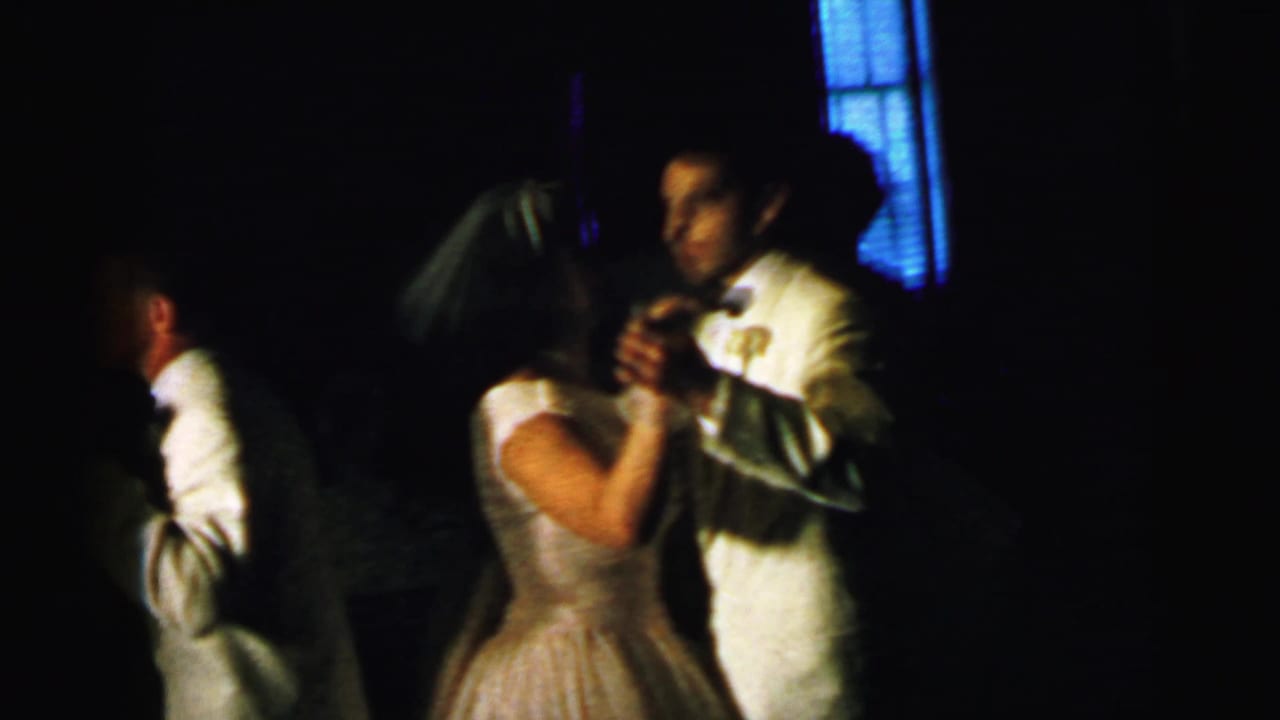 The bride and groom dance together in a dark reception interior with blue light visible through window blinds.