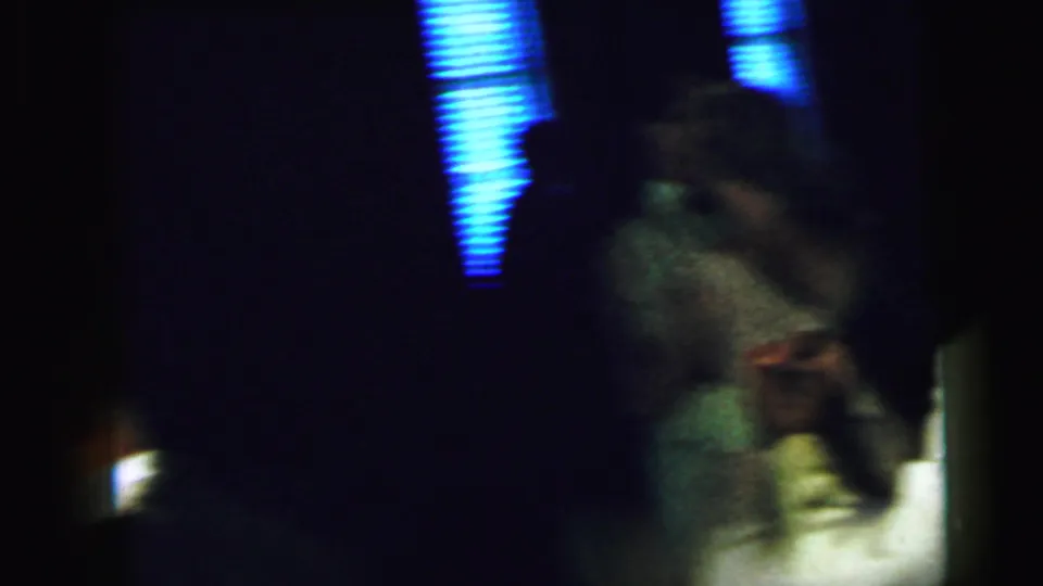 The couple remains in motion near blue-lit window blinds, with the bride's veil and the groom's light jacket partly visible.