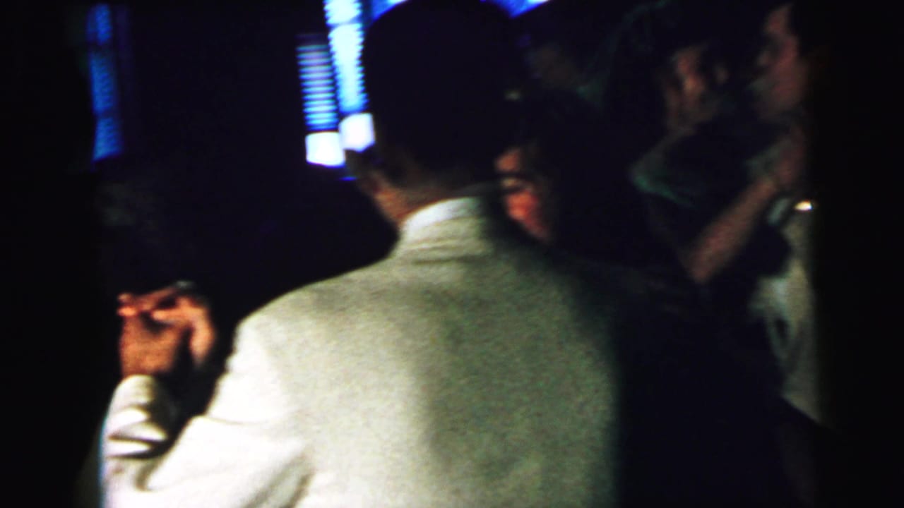 In this 1964 Camden home-movie frame, adults in formal attire dance closely in a dim interior with blue-lit blinds behind them.