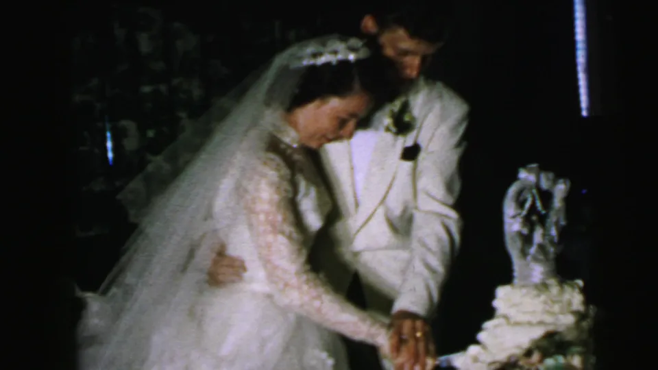 A bride in a veil and groom in a light jacket continue the cake-cutting ritual at a Camden wedding reception in 1964.