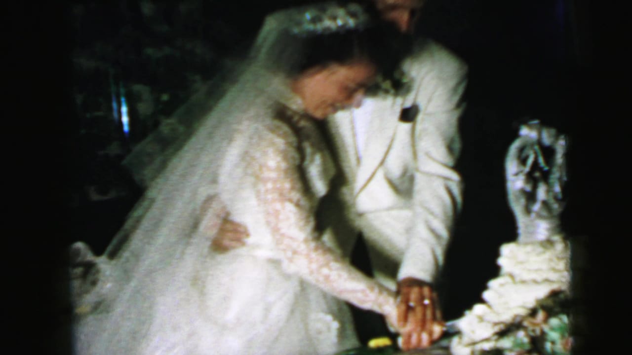 In 1964 Camden wedding footage, the bride and groom lean toward a tiered cake as their hands meet on the knife.