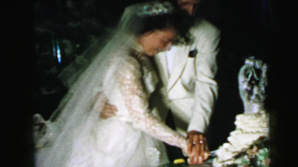 In 1964 Camden wedding footage, the bride and groom lean toward a tiered cake as their hands meet on the knife.