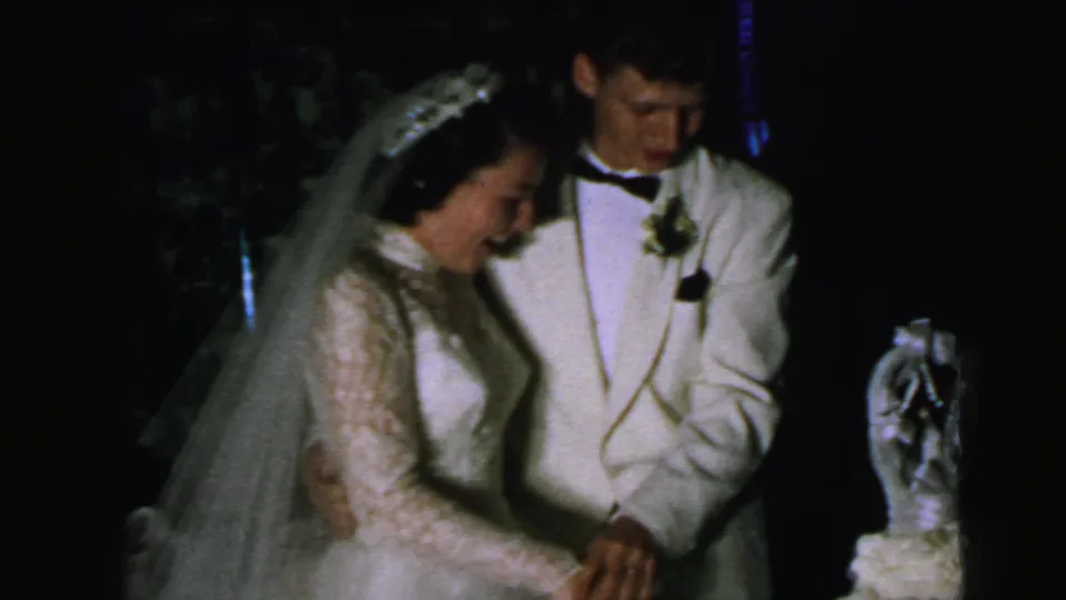 The bride in a lace gown and veil stands with the groom in a white jacket beside the cake during the 1964 Camden reception.
