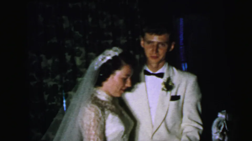 The couple stands near the decorated cake, with the groom facing the camera and the bride turned slightly downward.