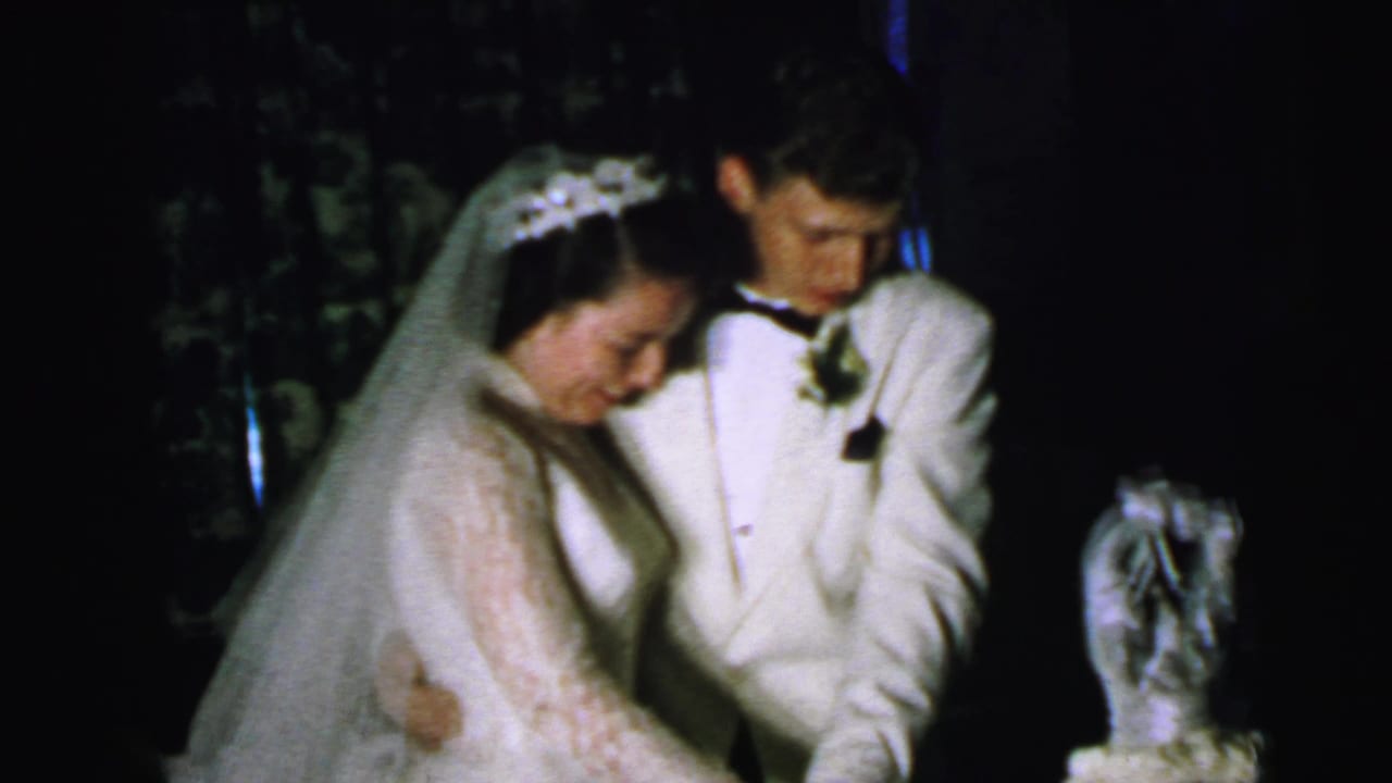 The bride and groom in formal wedding attire look down at the cake during a 1964 home-movie reception scene in Camden.