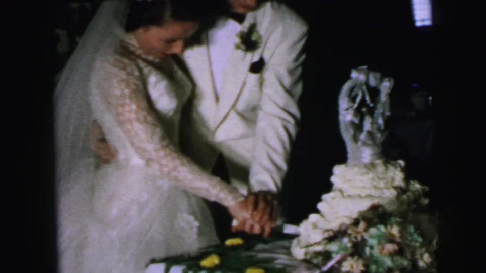 The bride and groom jointly press a knife into the decorated wedding cake in this Camden, New Jersey home-movie frame.