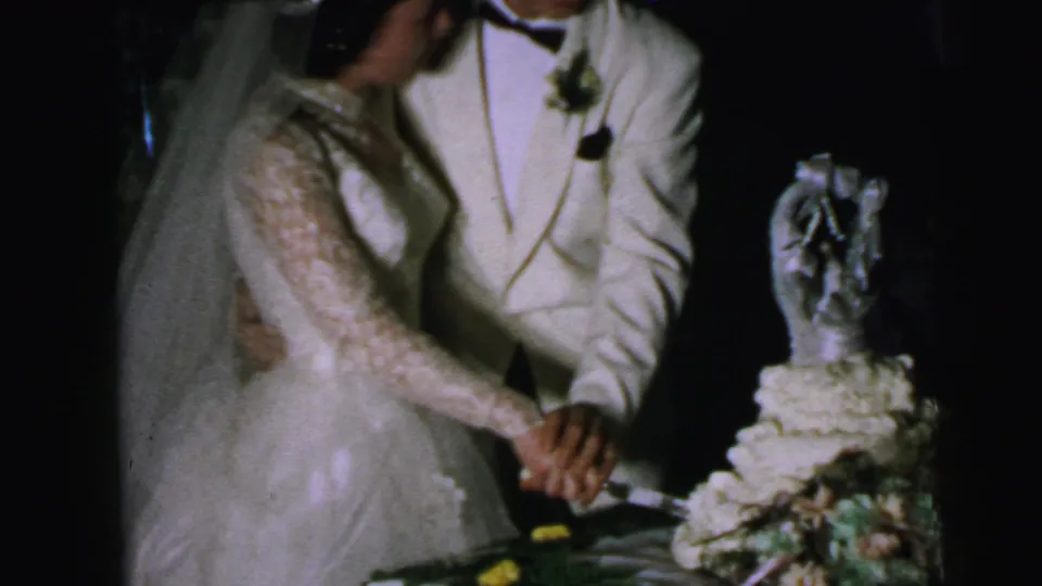 The bride leans over the cake with the groom close behind, continuing the wedding cake-cutting scene in 1964 Camden.