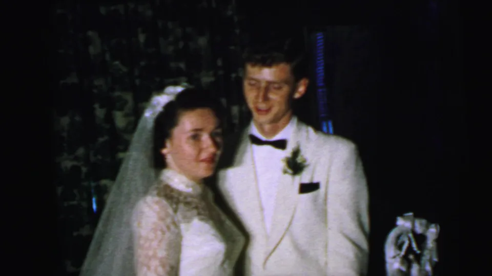 The bride and groom stand side by side near the cake, briefly facing the home-movie camera during the reception.