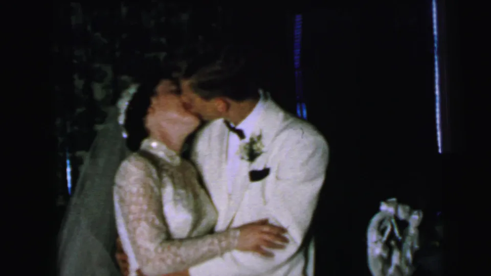 The groom leans in to kiss the bride beside the tiered cake in this 1964 Camden wedding reception footage.