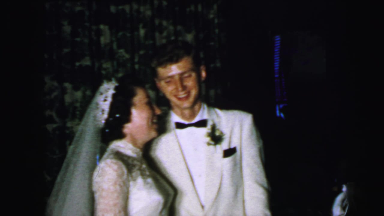 The bride turns toward the groom in this 1964 Camden wedding home-movie frame, with both figures standing before dark curtains.