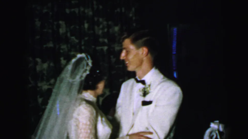 The bride stands close to the groom during the 1964 Camden wedding scene, with his white jacket and her veil prominent against the dark backdrop.