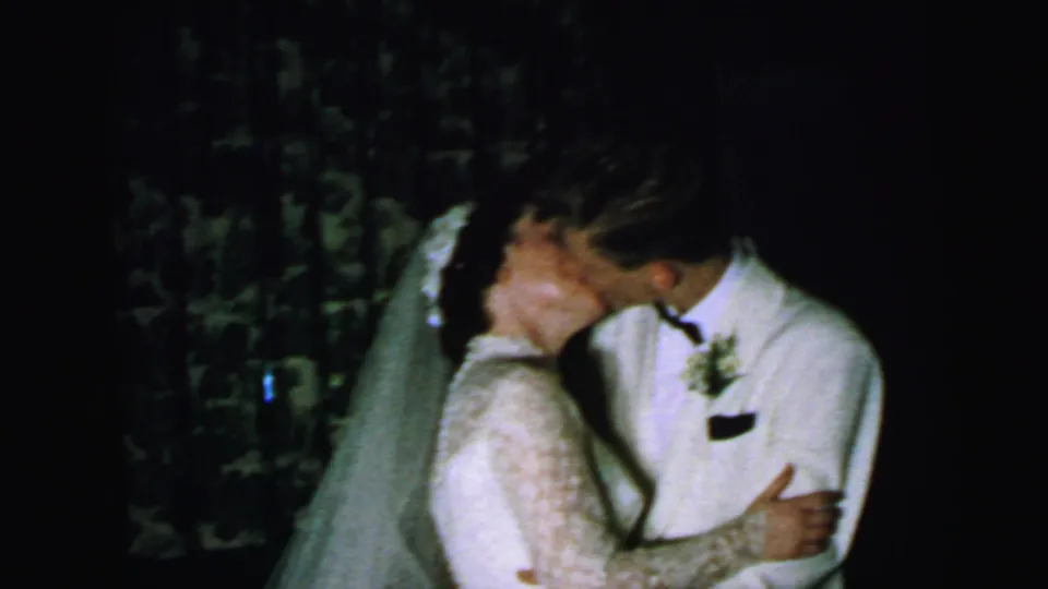 The bride and groom share a kiss in the supported 1964 Camden wedding footage, framed as an intimate home-movie reception moment.