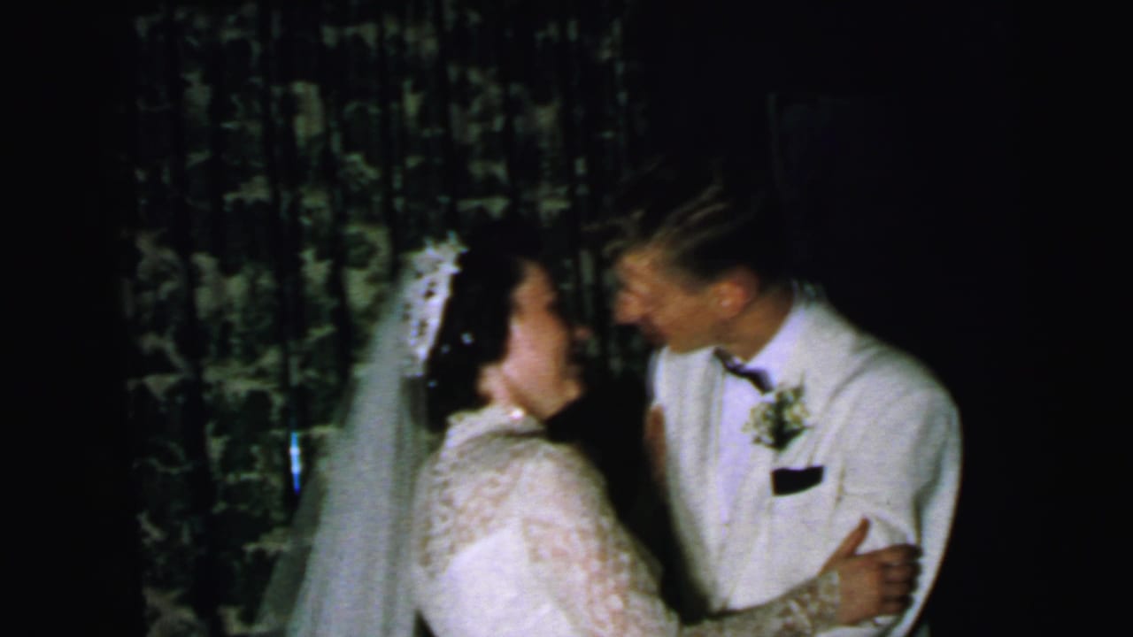 Bride in a veil and groom in a white tuxedo kiss indoors beside patterned curtains.