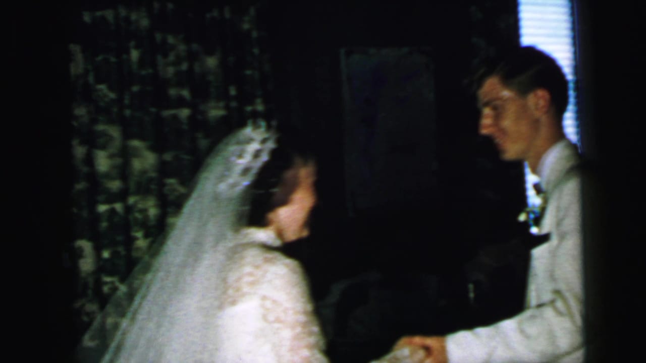 Bride and groom face each other indoors, with the groom reaching toward her hand.