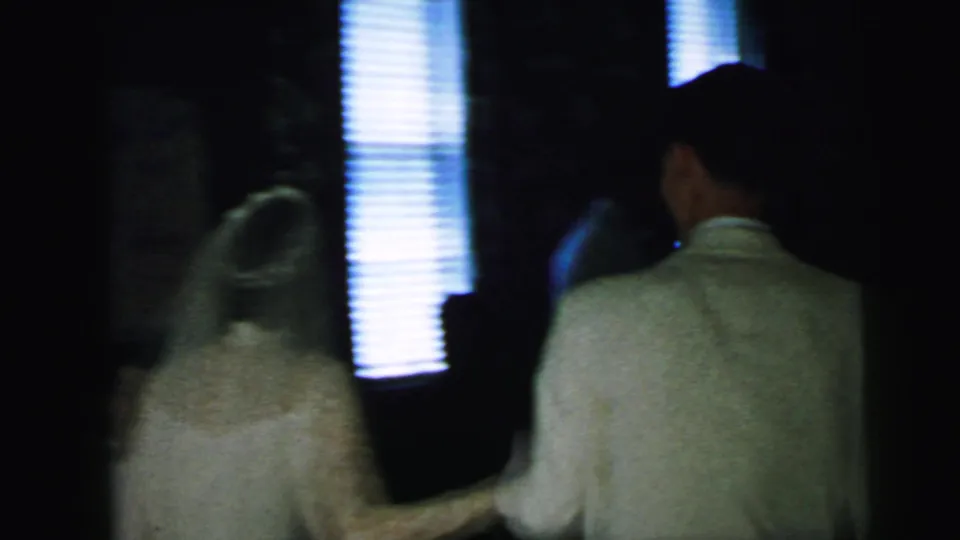 The bride and groom are seen from behind walking together in a dark private interior with bright window blinds.