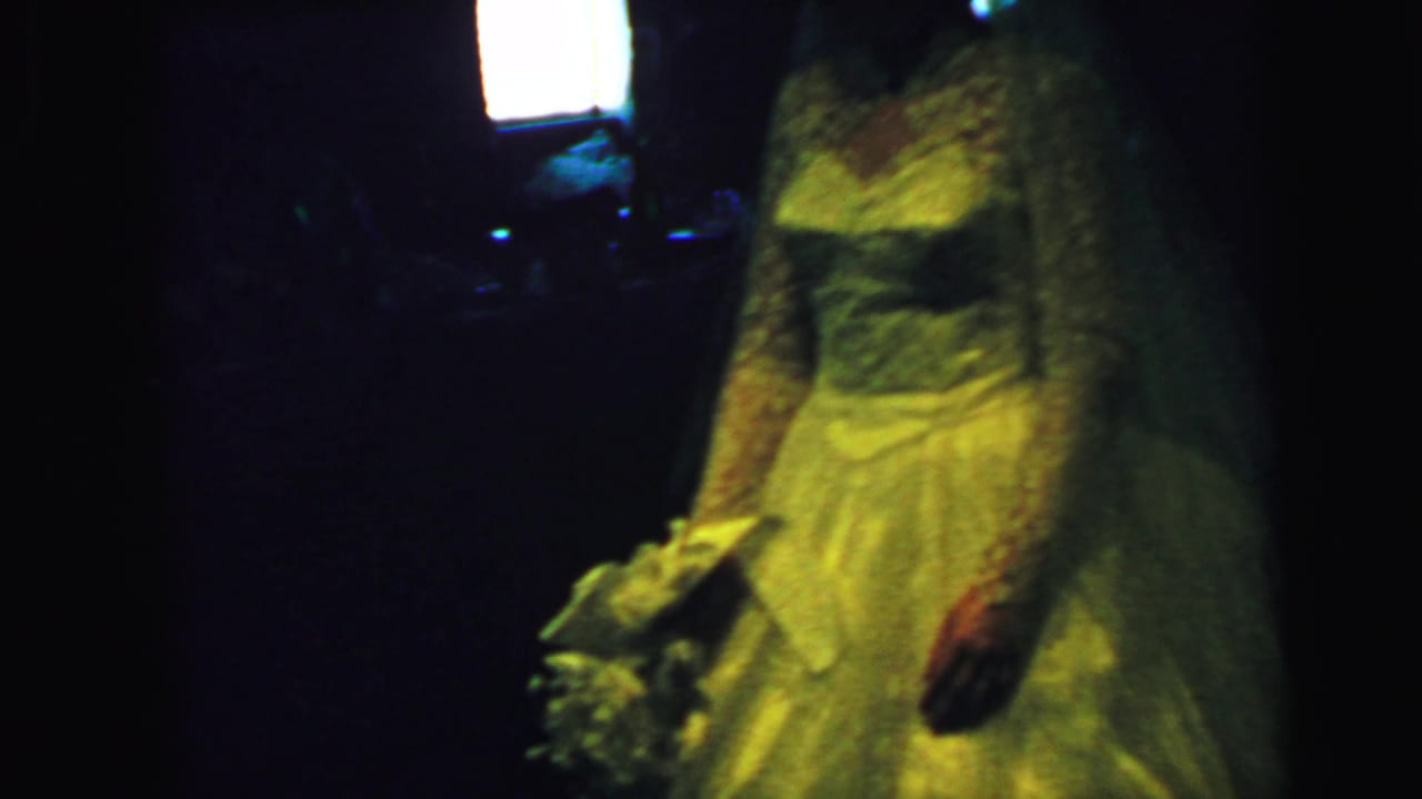 A bride in a 1960s white lace wedding gown stands in a dim Camden, New Jersey interior with a bouquet partly visible.