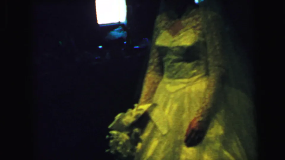 A bride in a 1960s white lace wedding gown stands in a dim Camden, New Jersey interior with a bouquet partly visible.