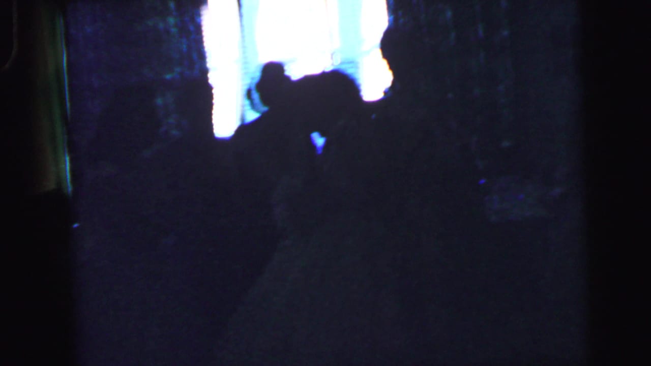 A dim 8mm frame from the Camden wedding reception shows guests reduced to silhouettes beside a bright window.