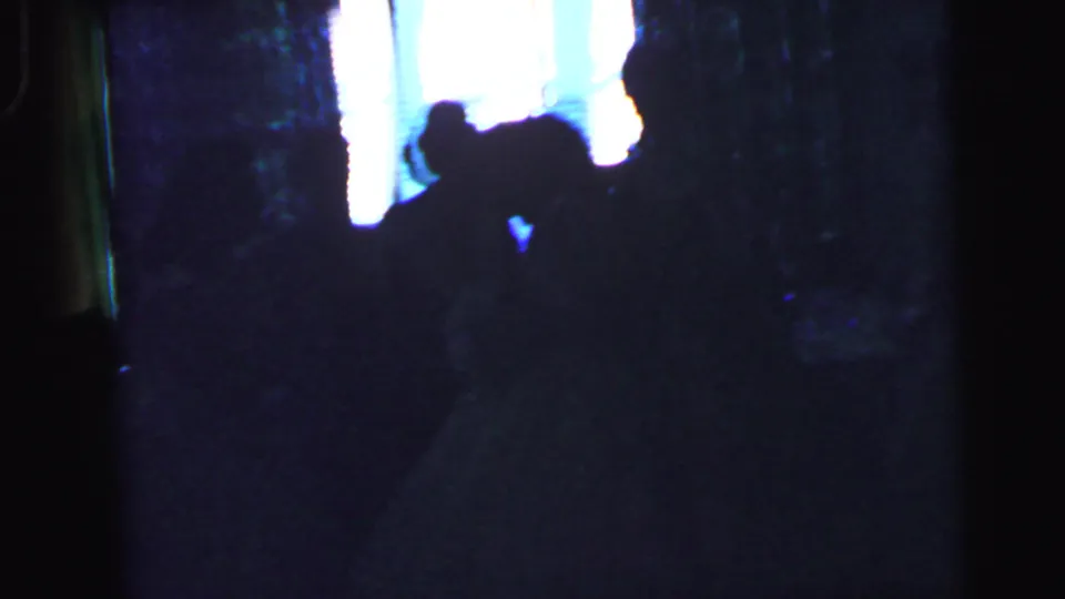 A dim 8mm frame from the Camden wedding reception shows guests reduced to silhouettes beside a bright window.