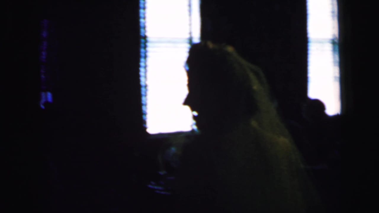 Silhouetted guest with long hair appears beside bright windows in a dark room.