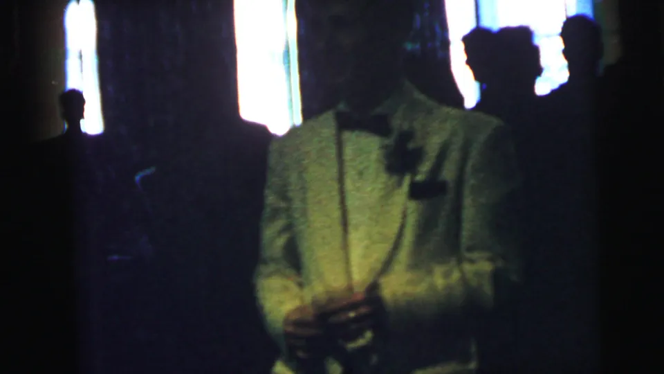 In a Camden, New Jersey 1964 home-movie frame, a man in a white tuxedo jacket stands backlit among shadowed guests.