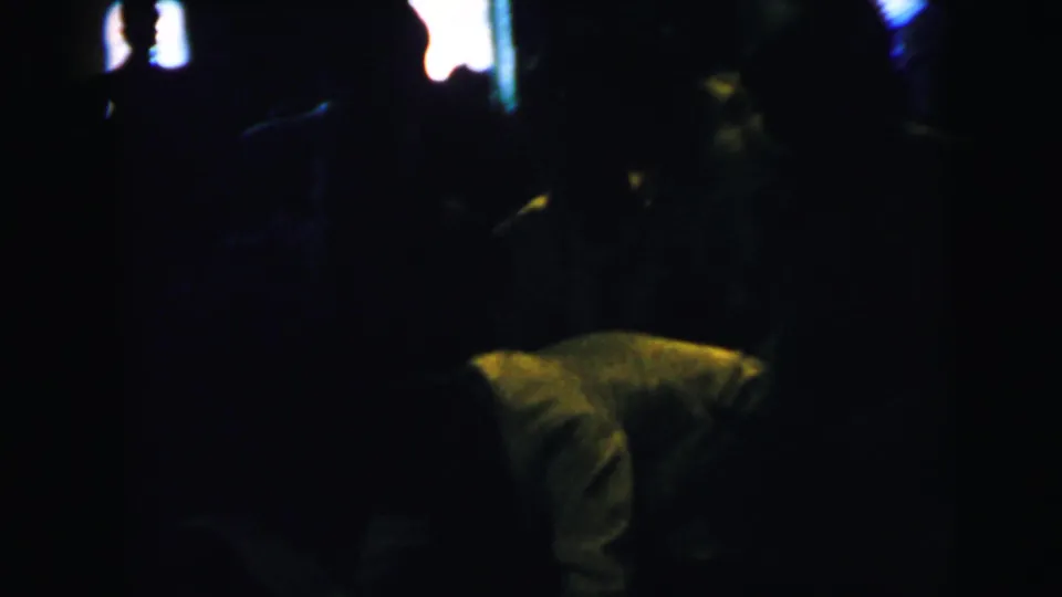 A dim, soft-focus home-movie frame shows shadowed guests in a formal interior, with a pale clothed figure low in the foreground.