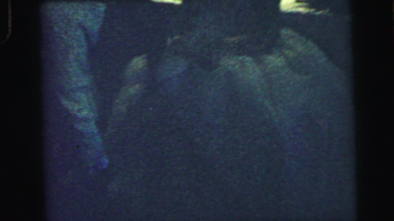 Dim 8mm frame of a suited man bending near a bride's white dress during a wedding garter moment.