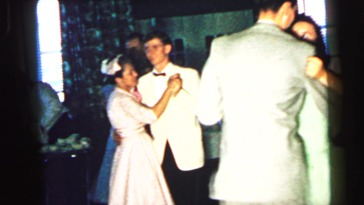 1964 Camden home-movie frame showing adults in suits and dresses dancing closely in a dim private interior.
