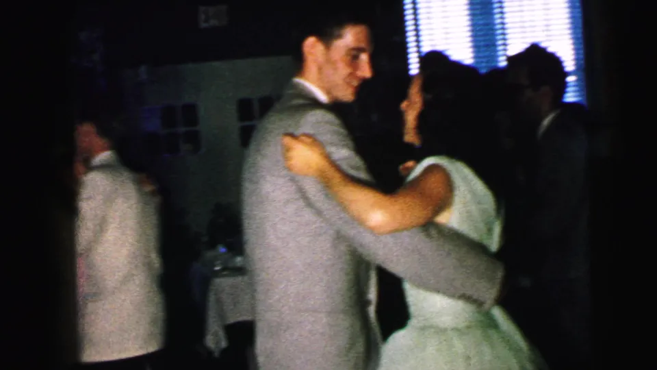 1964 Camden home-movie frame of a couple dancing close together in a dark interior, with other guests partly visible.