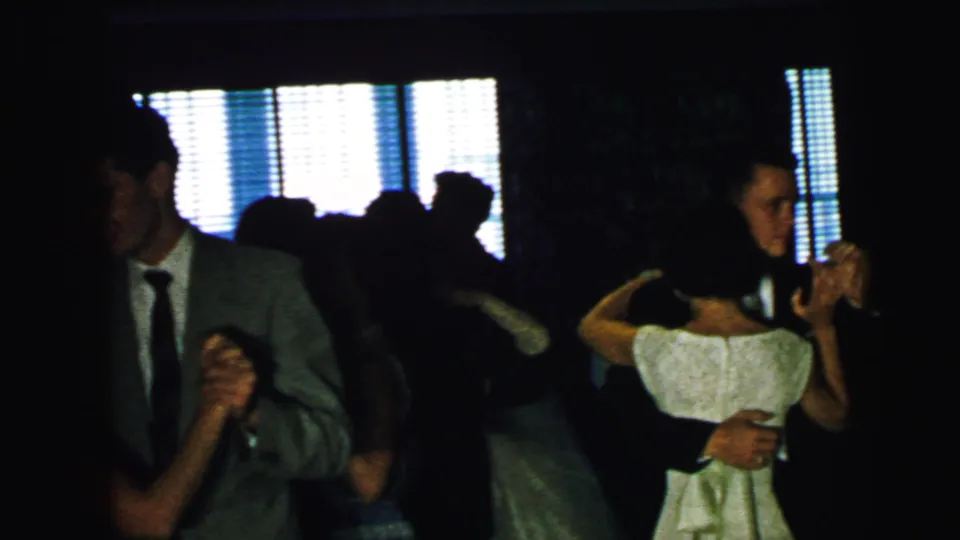 1964 Camden home-movie frame showing adults in suits and dresses dancing in a dark interior against bright window blinds.
