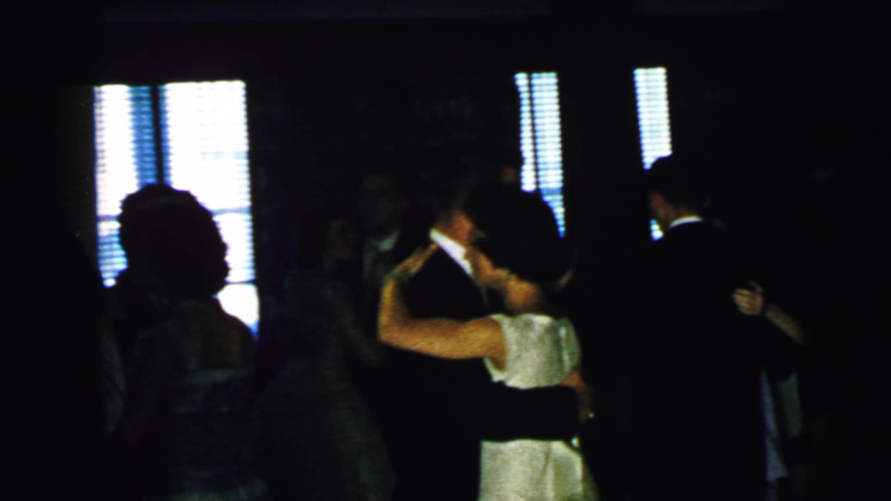 Several couples dance in silhouette near bright windows with blinds.
