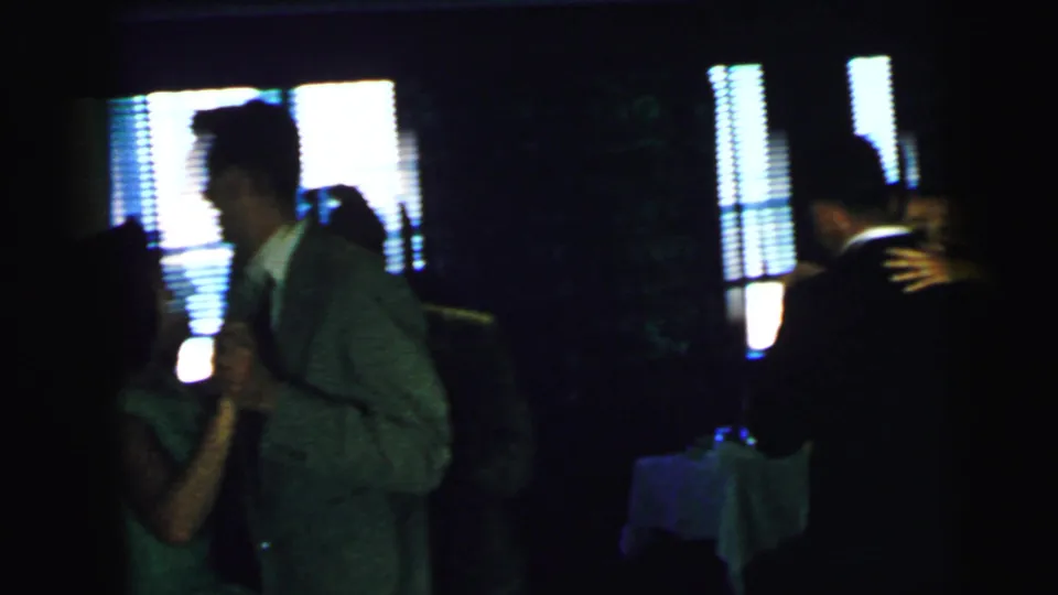 A handheld 8mm frame shows a suited man dancing near a woman in a dim Camden party interior, with window blinds glowing behind them.