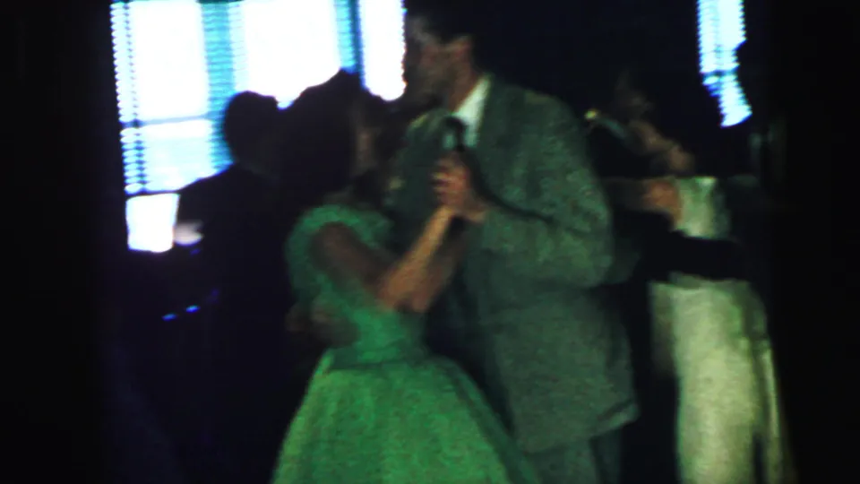 A woman in a green dress and a tall suited man dance in a dim 1964 Camden party scene, with another couple partly visible behind them.