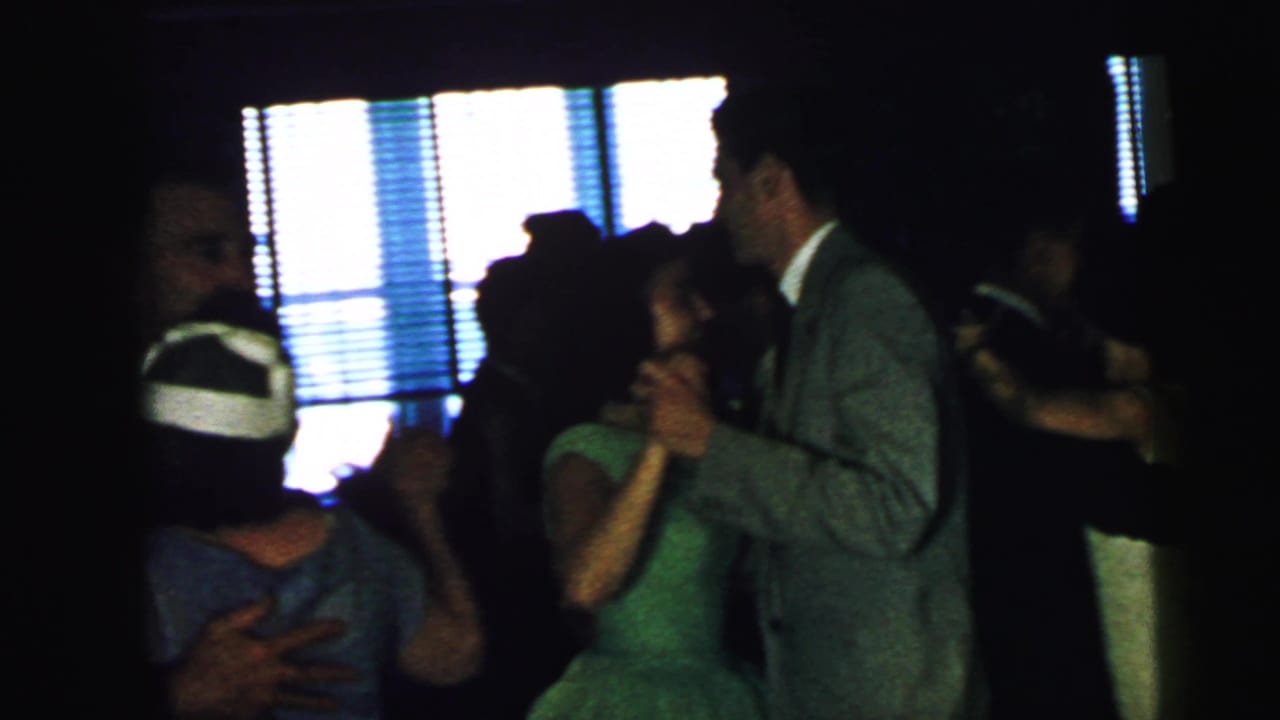 At a 1964 Camden wedding reception, several guests dance closely in a dark hall, including a woman in a green dress and a man in a gray suit.
