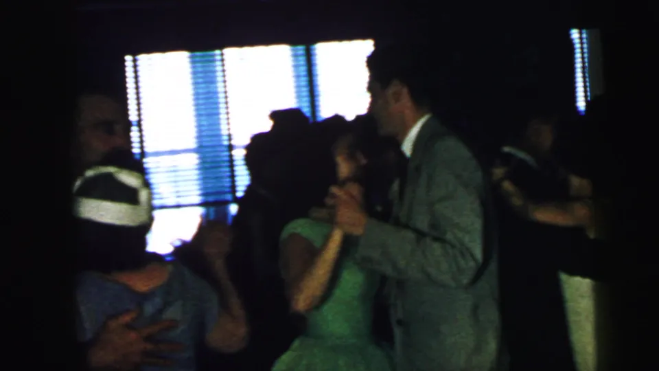 At a 1964 Camden wedding reception, several guests dance closely in a dark hall, including a woman in a green dress and a man in a gray suit.