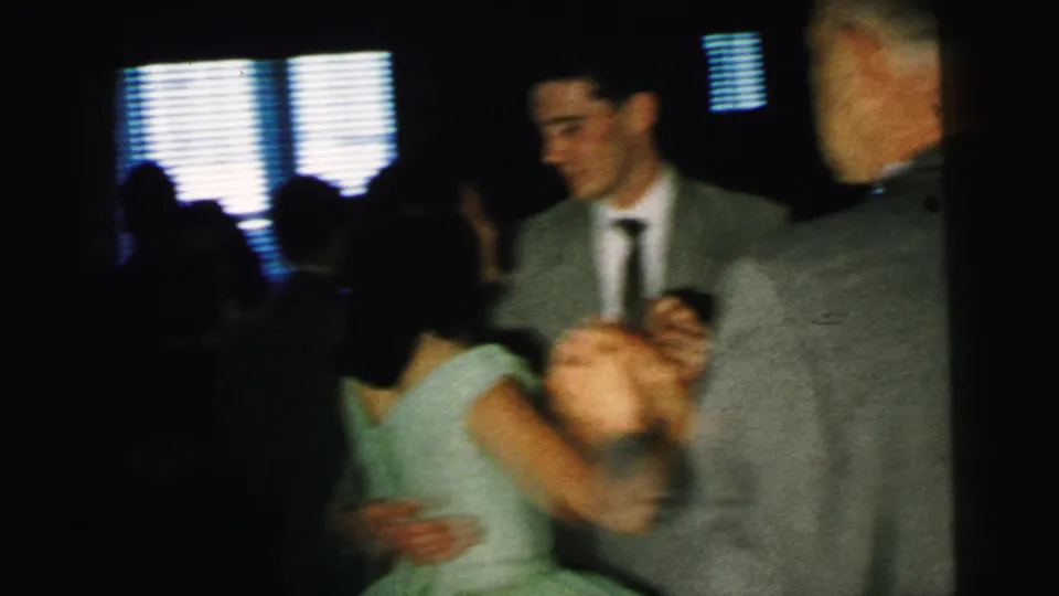 A woman in a pale dress dances with a partner as another suited guest appears beside them in the dim 1964 Camden reception footage.