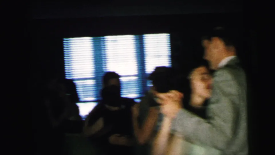A man in a light suit dances with a woman near bright blinds in the Camden wedding reception home movie.