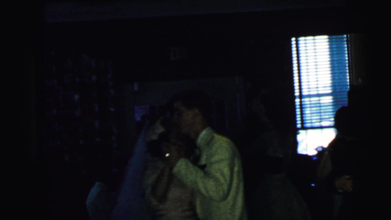 1964 Camden home-movie frame showing a couple dancing in a dark interior near window blinds and an EXIT sign.
