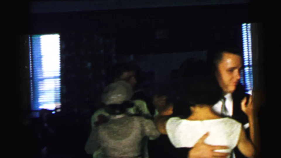 1964 Camden home-movie frame with multiple couples dancing indoors and a suited man visible near the camera at right.