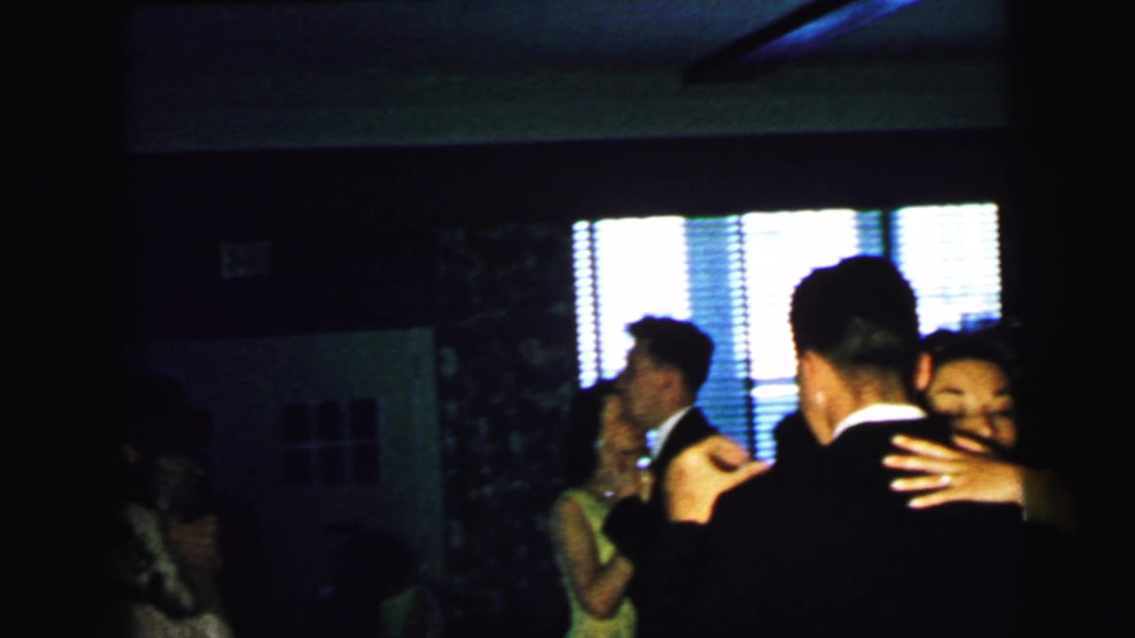 Couples in formal clothes dance in a dark room with bright window blinds behind them.