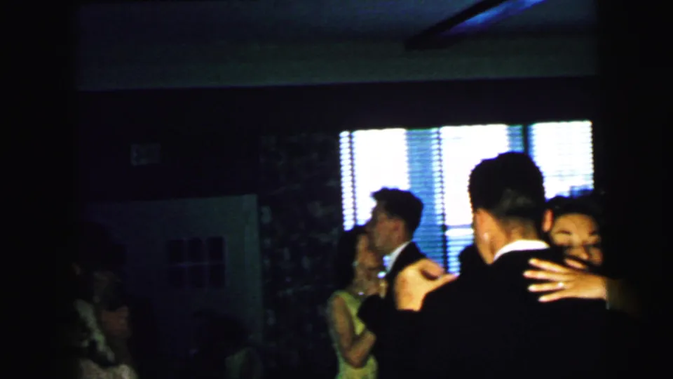 1964 Camden home-movie frame showing wedding guests dancing indoors, with formal clothing and bright blinds in the background.