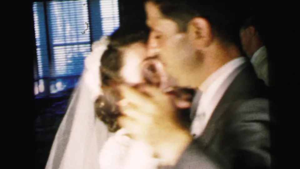 Bride and groom appear in close-up as they dance at the 1964 Camden reception, with guests and window blinds behind them.