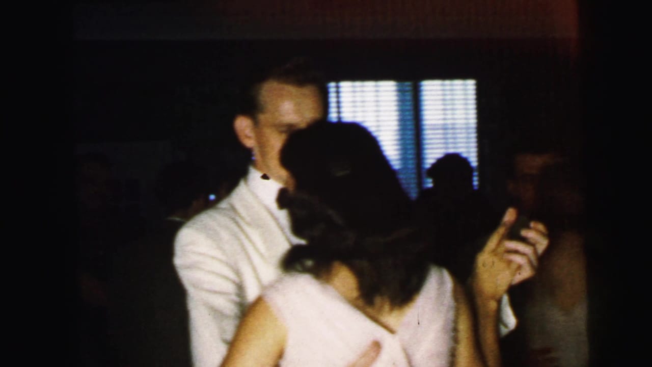 A man in a white dinner jacket dances with a woman in a light dress in a dim room.