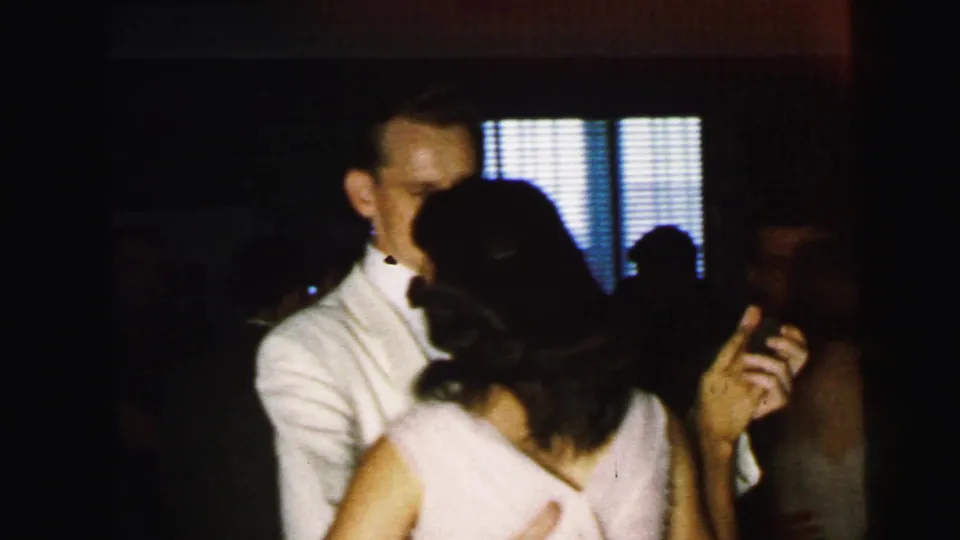 1964 Camden home-movie frame showing a formal-dressed couple dancing indoors, with other guests and vertical blinds behind them.